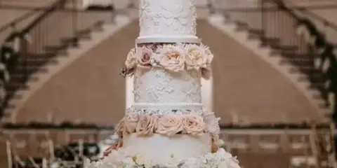 A tall, multi-tiered white wedding cake decorated with floral arrangements and fondant flowers, displayed in a large event hall.