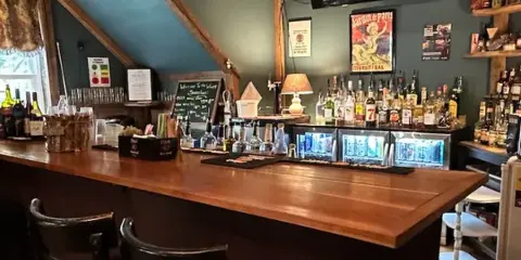 Interior view of a wooden bar counter with shelves of liquor bottles, glassware, and bar equipment in a lodge setting.
