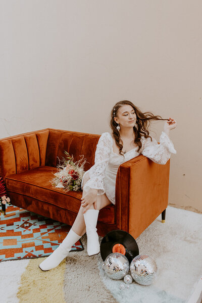 A bride in a white lace dress sits on a burnt orange velvet couch, twirling her hair.