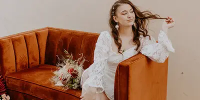A bride in a white lace dress sits on a burnt orange velvet couch, twirling her hair.