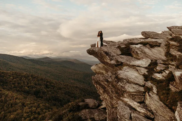 A bride and groom embracing on a rocky mountain cliff overlooking a forested valley.