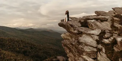 A bride and groom embracing on a rocky mountain cliff overlooking a forested valley.
