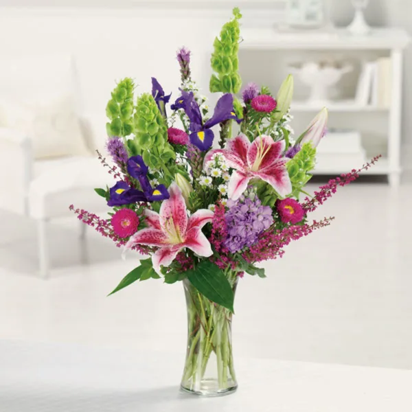 A tall mixed bouquet of lilies, irises, and purple flowers in a clear glass vase