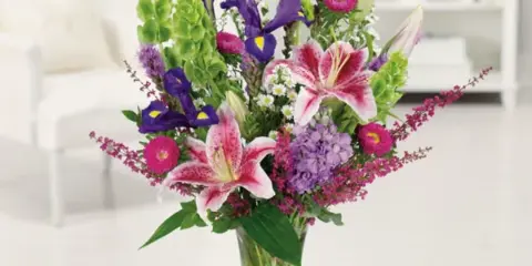 A tall mixed bouquet of lilies, irises, and purple flowers in a clear glass vase
