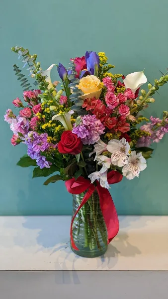 A colorful floral arrangement featuring roses, calla lilies, and alstroemeria in a glass vase with a red ribbon.
