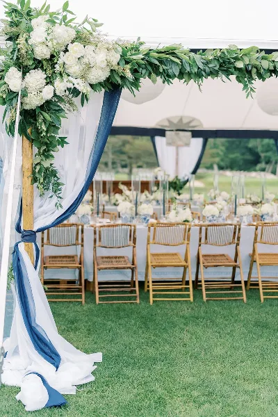 A wedding arch decorated with white flowers and greenery, with blue and white drapery, set up inside a large white tent with a row of wooden chairs and a long dining table in the background.