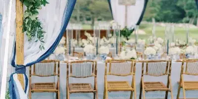 A wedding arch decorated with white flowers and greenery, with blue and white drapery, set up inside a large white tent with a row of wooden chairs and a long dining table in the background.