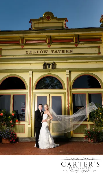 Bride and groom posing in front of the Yellow Tavern building with the bride’s veil blowing in the wind.