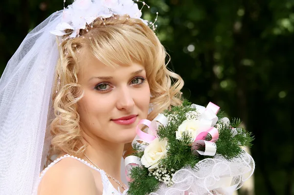 A bride with styled blonde hair, veil, and makeup holding a bouquet of white roses and greenery.