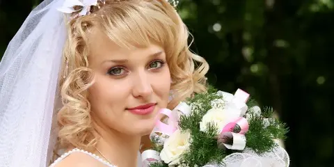 A bride with styled blonde hair, veil, and makeup holding a bouquet of white roses and greenery.