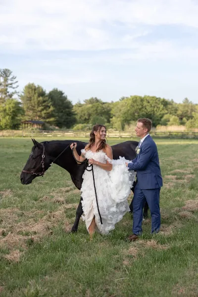 A bride and groom standing in a grassy field with a black horse, representing an equestrian wedding venue.