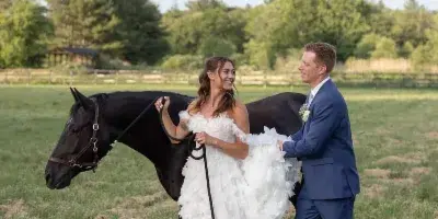 A bride and groom standing in a grassy field with a black horse, representing an equestrian wedding venue.