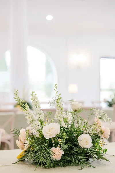 A detailed wedding floral centerpiece featuring white roses, pale pink carnations, delphiniums, and lush greenery on a table.