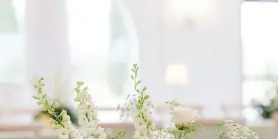 A detailed wedding floral centerpiece featuring white roses, pale pink carnations, delphiniums, and lush greenery on a table.