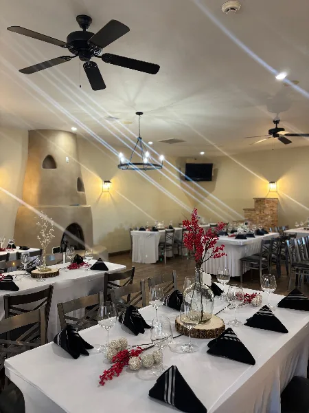 Interior view of a banquet hall with multiple tables set for an event, featuring white tablecloths, black napkins, and a large adobe fireplace in the background.