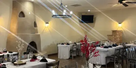 Interior view of a banquet hall with multiple tables set for an event, featuring white tablecloths, black napkins, and a large adobe fireplace in the background.