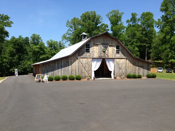 Rustic wooden barn with white drapery at the entrance, surrounded by trees and a paved driveway.