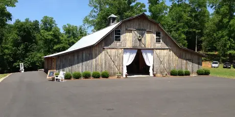 Rustic wooden barn with white drapery at the entrance, surrounded by trees and a paved driveway.