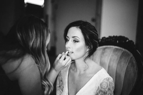 A makeup artist applies lipstick to a seated bride with an updo hairstyle.