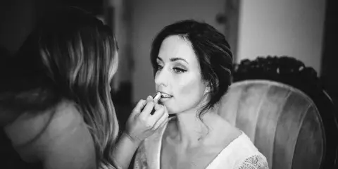 A makeup artist applies lipstick to a seated bride with an updo hairstyle.