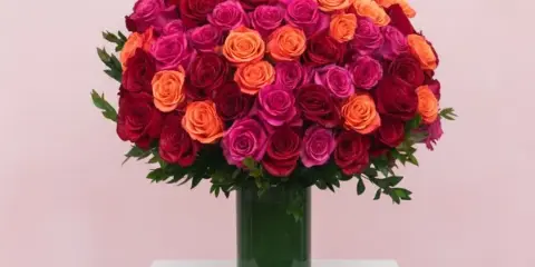 A round bouquet of red, pink, and orange roses in a green glass vase.
