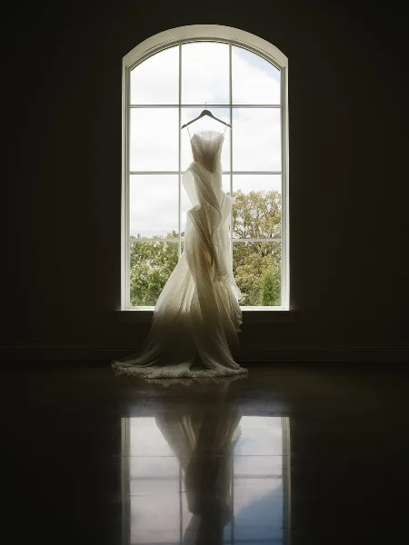 A white wedding dress hanging on a hanger in front of a large arched window with a view of trees outside.