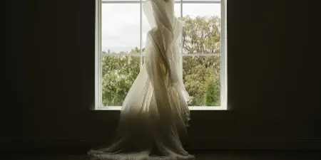 A white wedding dress hanging on a hanger in front of a large arched window with a view of trees outside.
