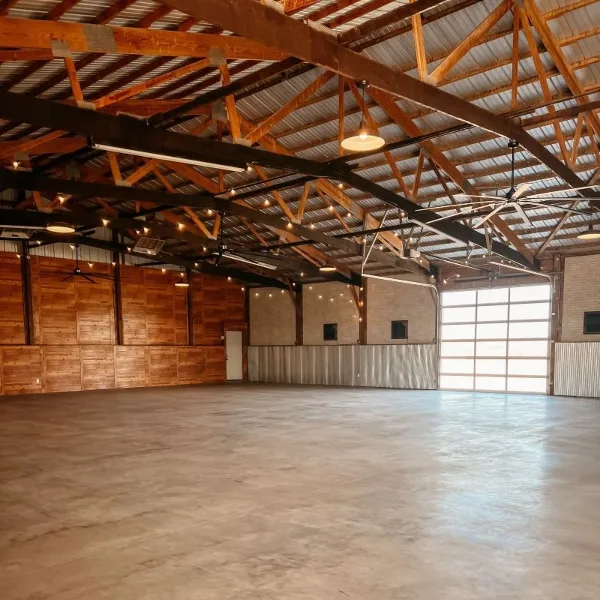 Interior view of a large event venue with exposed wooden roof trusses, string lights, and a polished concrete floor.