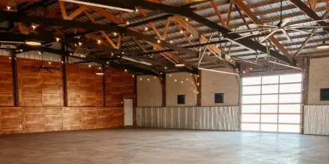 Interior view of a large event venue with exposed wooden roof trusses, string lights, and a polished concrete floor.