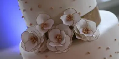 A close-up of a white wedding cake tier decorated with white sugar flowers and a burlap ribbon.