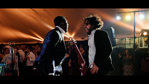 Cinematic still of a live band performing at a wedding reception with guests in the background.