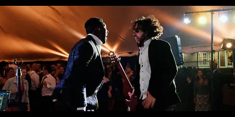 Cinematic still of a live band performing at a wedding reception with guests in the background.