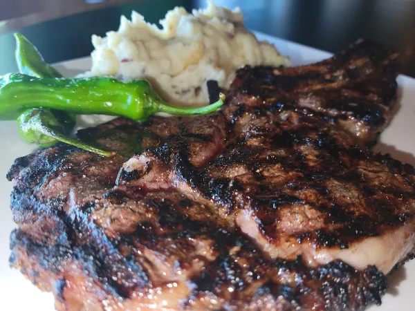 Grilled steak with charred crust served with mashed potatoes and green peppers on a white plate.