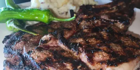Grilled steak with charred crust served with mashed potatoes and green peppers on a white plate.