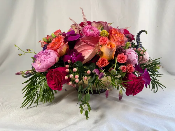 A colorful floral arrangement featuring roses, peonies, orchids, and greenery in a vase on a white background.