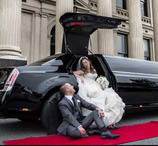 A bride and groom posing with a black limousine on a red carpet.
