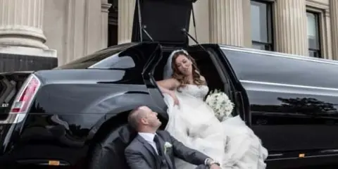 A bride and groom posing with a black limousine on a red carpet.