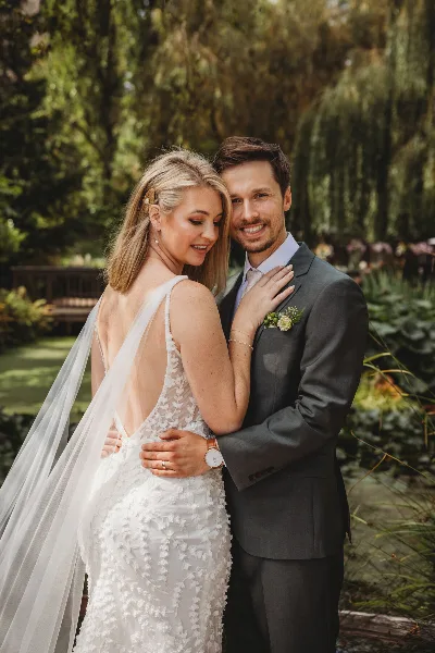 A bride and groom embracing in a garden setting, with the bride wearing a white lace dress and the groom in a grey suit.