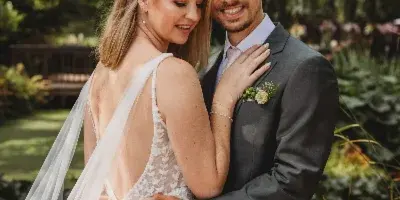 A bride and groom embracing in a garden setting, with the bride wearing a white lace dress and the groom in a grey suit.