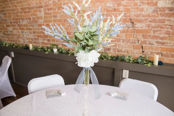 A wedding table setting with a floral centerpiece and white chairs in front of a brick wall.