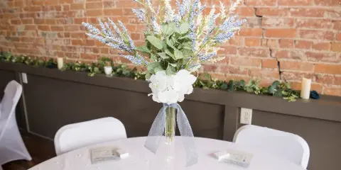 A wedding table setting with a floral centerpiece and white chairs in front of a brick wall.