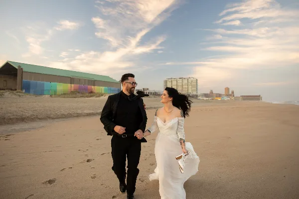 A bride and groom walking hand-in-hand on a sandy beach with a colorful building and city skyline in the background.