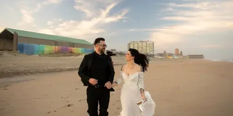 A bride and groom walking hand-in-hand on a sandy beach with a colorful building and city skyline in the background.