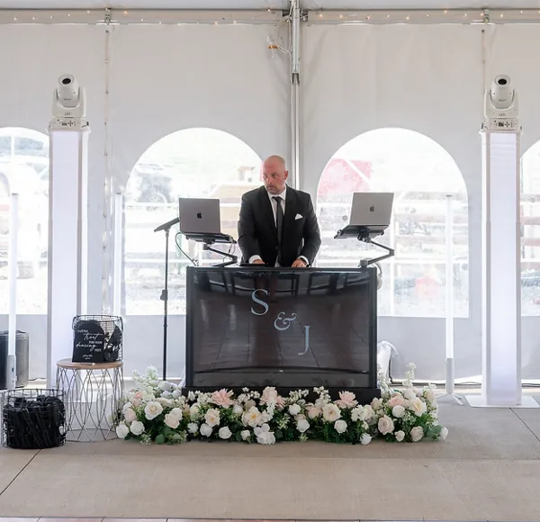 A professional DJ in a suit operating a customized booth with a monogram screen and laptops at a wedding reception.