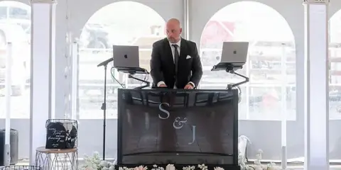 A professional DJ in a suit operating a customized booth with a monogram screen and laptops at a wedding reception.
