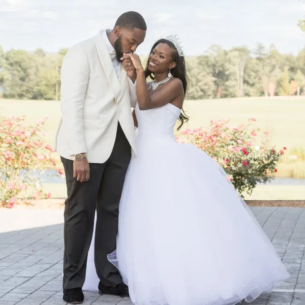 A bride in a white gown and a groom in a white jacket posing together outdoors.