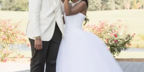 A bride in a white gown and a groom in a white jacket posing together outdoors.
