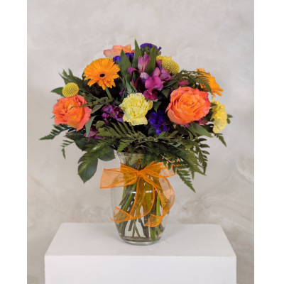 A colorful floral arrangement featuring orange roses, yellow carnations, and greenery in a clear glass vase tied with an orange ribbon.