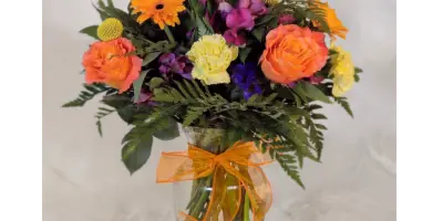 A colorful floral arrangement featuring orange roses, yellow carnations, and greenery in a clear glass vase tied with an orange ribbon.
