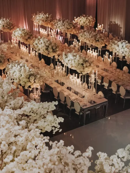 A wedding reception hall featuring long banquet tables with white floral centerpieces and tall, elevated floral installations with taper candles.